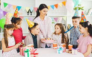 Mother holding a cake and kid blowing out candles. 5 other kids wearing party hats around the cake watching.