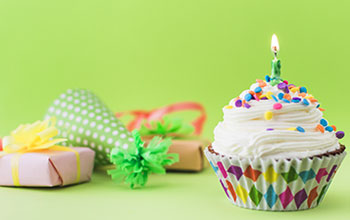 Cupcake with 1 candle, wrapped present, and a party hat all on a green table with a green background.